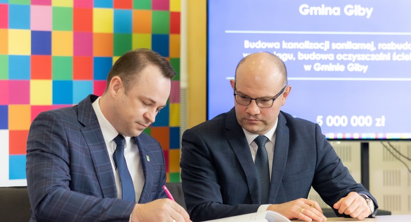Zdjęcie dwóch mężczyzn siedzących za stołem, jeden z nich podpisuje dokument
