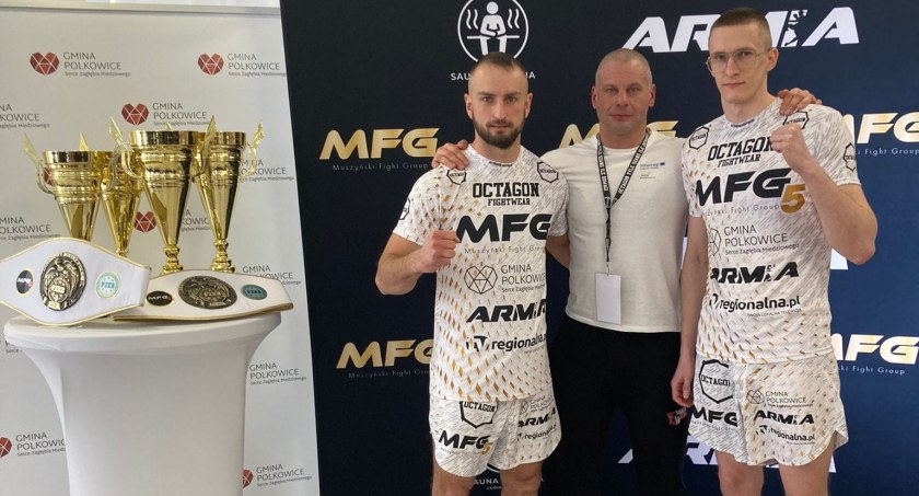 Zdjęcie trzech mężczyzn przy stole z trofeami na tle ścianki z logo