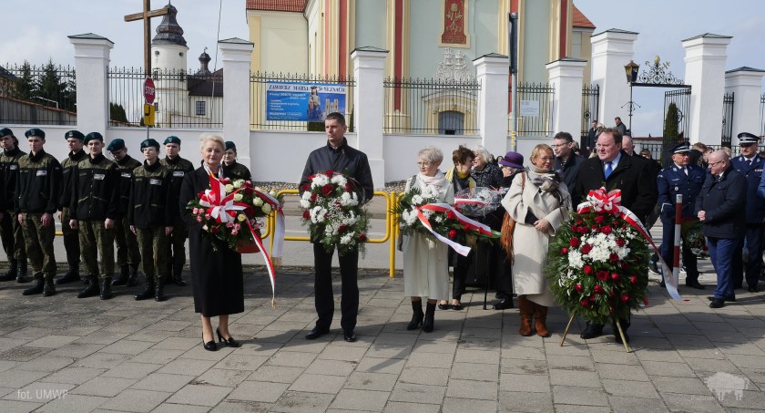 Zdjęcie grupy osób z wieńcami i wiązankami kwiatów, stojących na placu św. Agaty z Bazyliką w tle