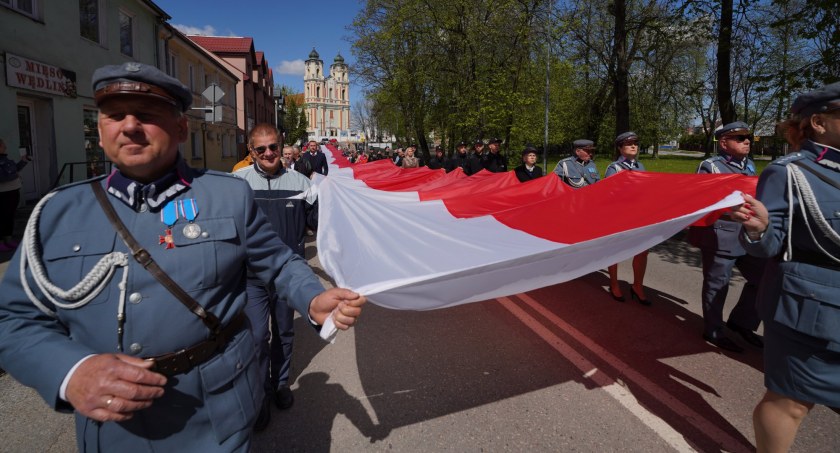 Zdjęcie osób w mundurach niosących długą flagę RP, maszerując jezdnią ul. Piłsudskiego