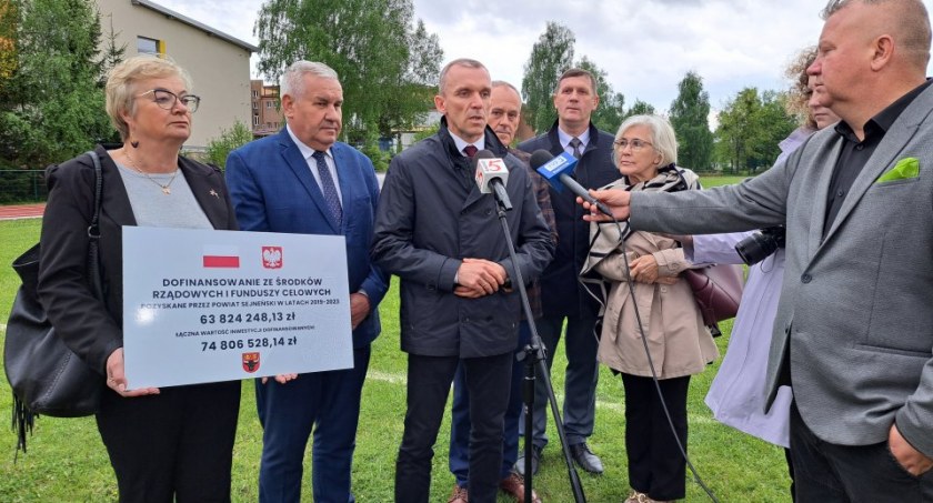 Zdjęcie osób stojących przed mikrofonami na tle budowy obiektu sportowego